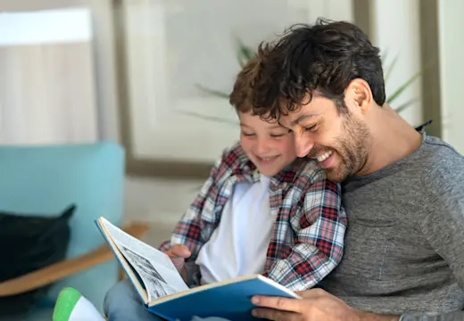 Father and son smiling while reading a book together at home.