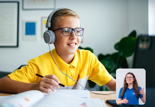 A cheerful young student with headphones participates in an online class, engaging with a tutor visible on the laptop screen, while sitting at a well-organised study area.
