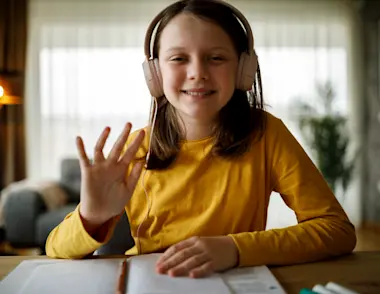 Young girl in a yellow top sitting at a computer with headphones waving to her online tutor.