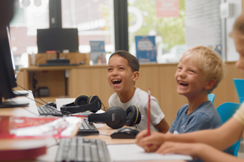 Two young boys sit next to each other at a desk laughing.